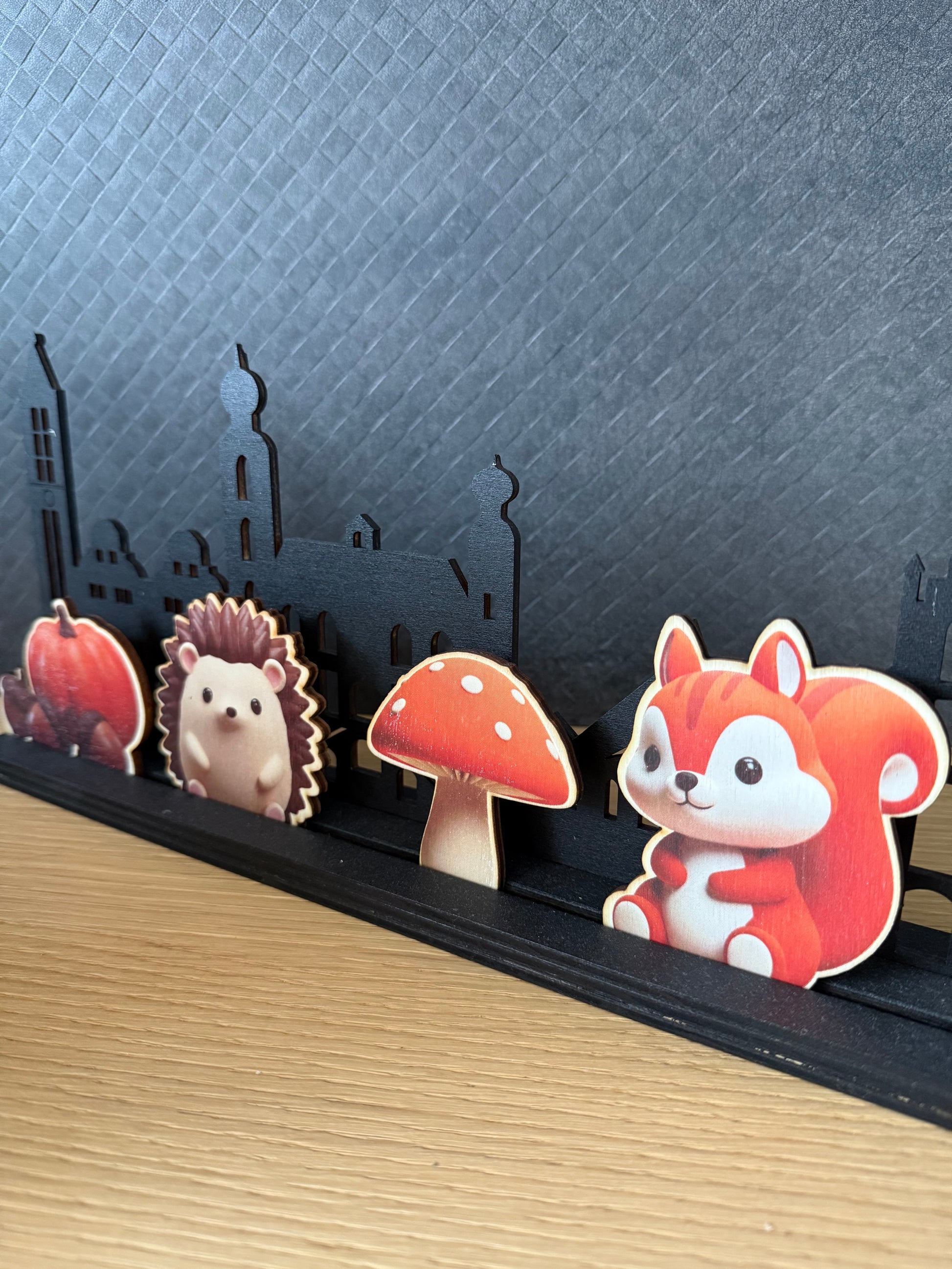 Een close-up van vier houten inzetstukjes met herfstthema (een pompoen met eikels, egel, paddenstoel en eekhoorn) geplaatst in het voetstuk van een zwarte, lasersneden skyline. De houten basis van de skyline en de tafel zijn zichtbaar, met een donkere, textuurrijke achtergrond.