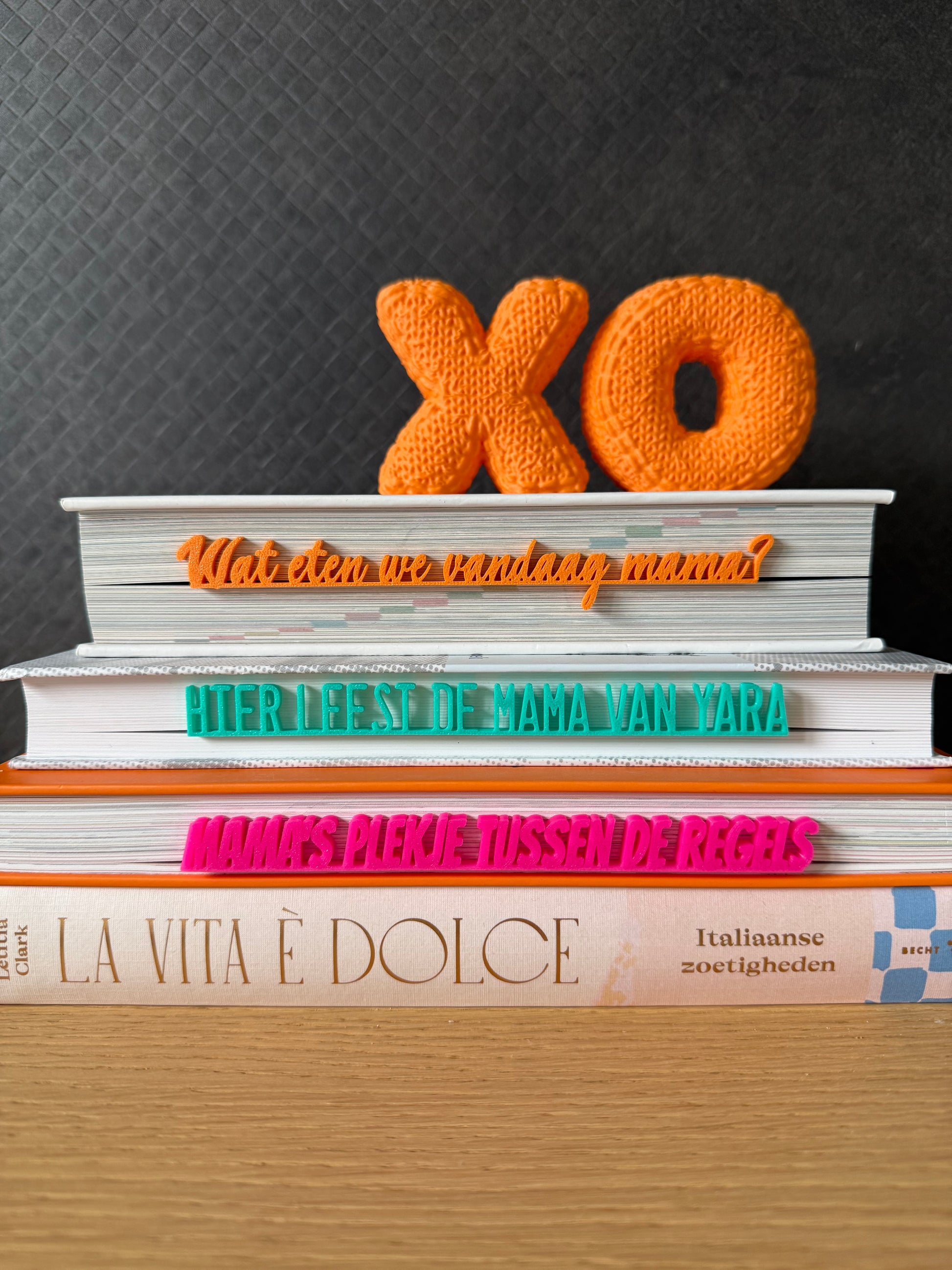 Drie 3D geprinte boekenleggers in oranje, turquoise en fuchsia op een stapel boeken, met teksten 'Wat eten we vandaag mama?', 'Hier leest de mama van Yara' en 'Mama's plekje tussen de regels'. Gemaakt door Traktatiecorner in Venlo.