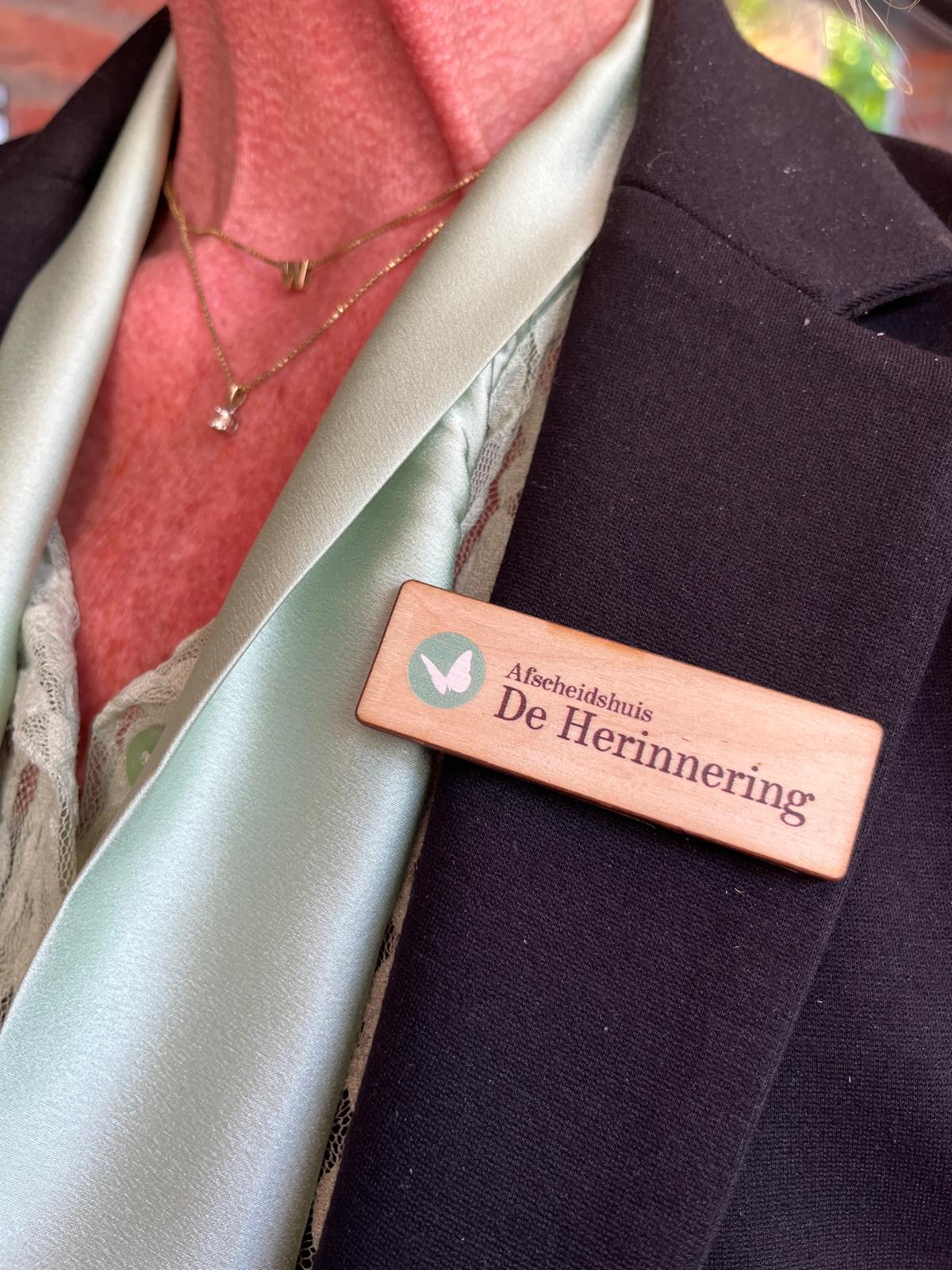 Houten badge met logo en naam van Afscheidshuis De Herinnering, gedragen op een zwarte jas met magneetsluiting