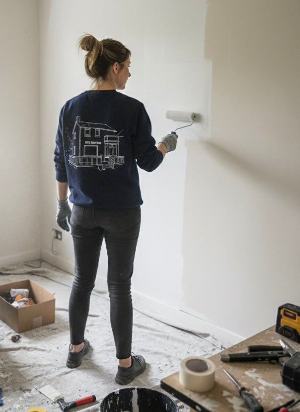 Een dame schildert een muur en draagt hierbij een sweater met lijntekening van het huis 